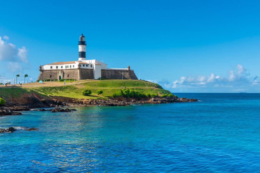 Vista do Farol da Barra em Salvador, com a praia ao redor e o mar azul ao fundo.
