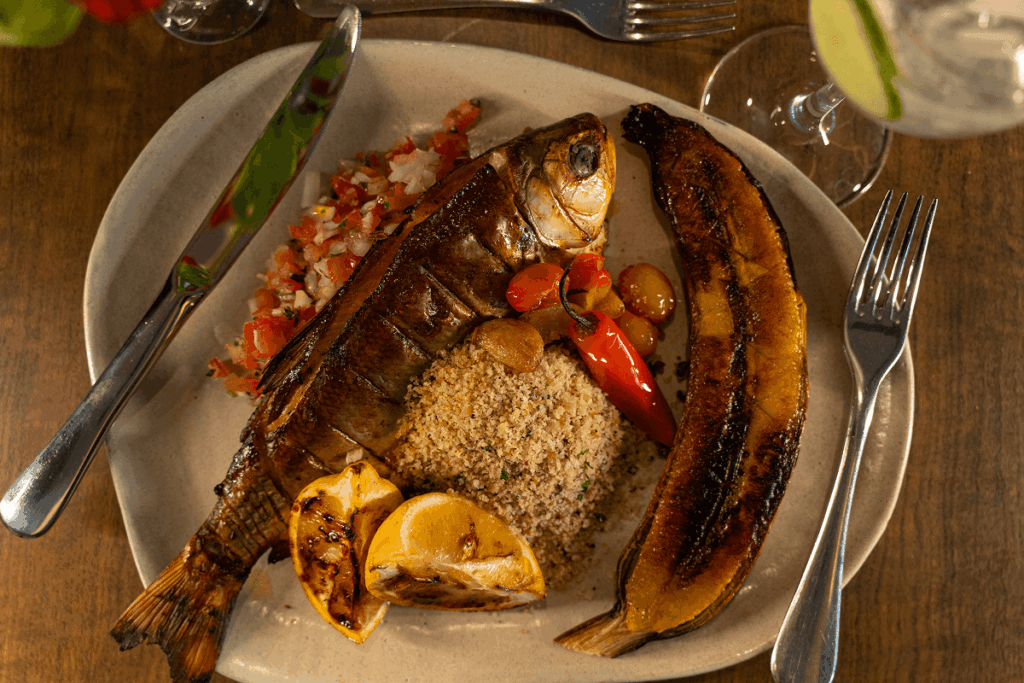 Prato escuro com um peixes inteiros grelhados com a pele tostada, guarnecidos com ervas frescas, grãos de pimenta-do-reino e fatias de limão.