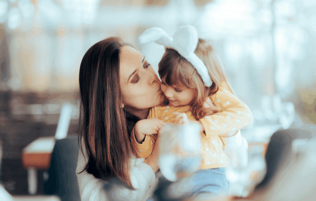 Uma mãe caucasiana está beijando sua filha, que usa orelhas de coelho de pelúcia. Juntas, elas celebram a Páscoa.