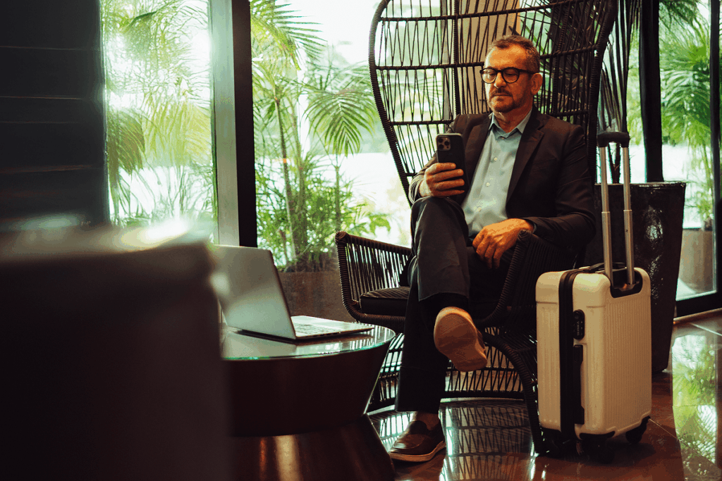 Um viajante de negócios de meia-idade com celular na mão sentado no lobby do hotel com uma mala.