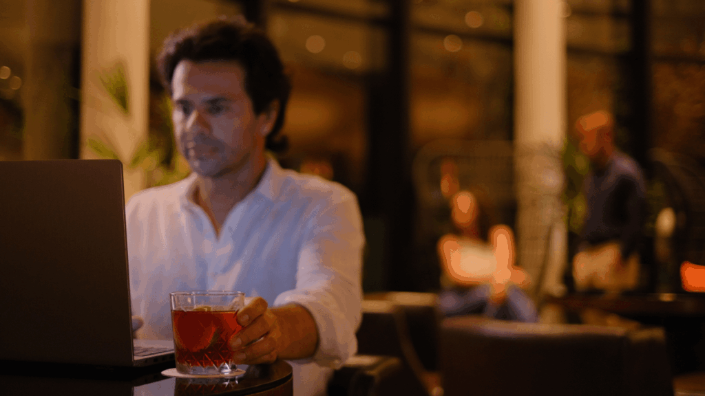 Homem caucasiano sentado em uma mesa, trabalhando em seu notebook em cima de uma e com uma bebida em sua mão. O espaço do loby é moderno e aconchegante.