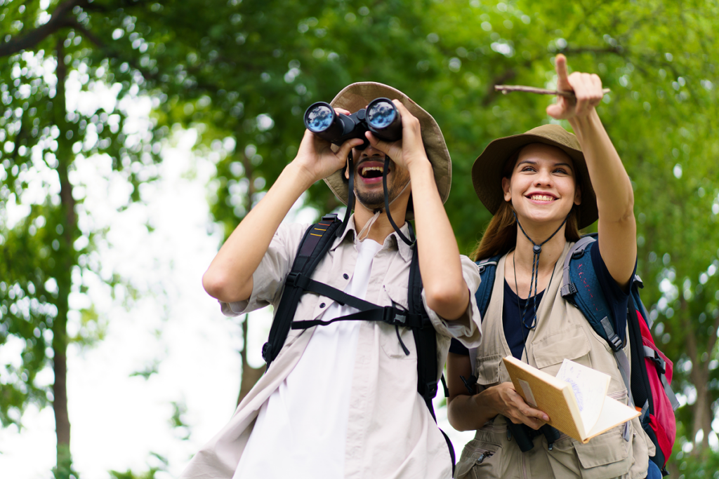Casal de homem e mulher caucasianos e excursionistas usa binóculos para observar pássaros na natureza, enquanto fazem anotações sobre eles.